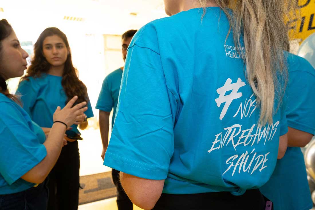 Membros da Corporate Heath em círculo, conversando de pé e com a camiseta azul da equipe com o seguinte dizer nas costas #Nós Entregamos Saúde.